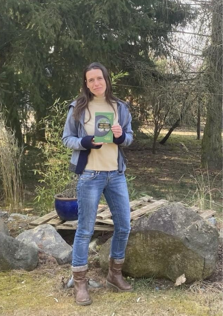 Kath stands outdoors on grass near large rocks and trees, holding her first book and facing the camera.